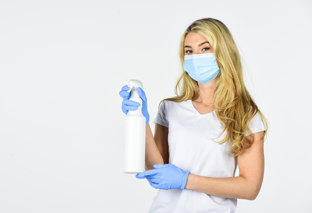 a young woman wearing a mask and gloves holding a spray bottle of cleaner