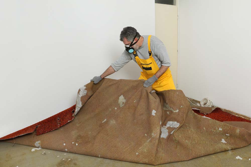 A man in yellow overalls and a gas mask removes a carpet from a room.