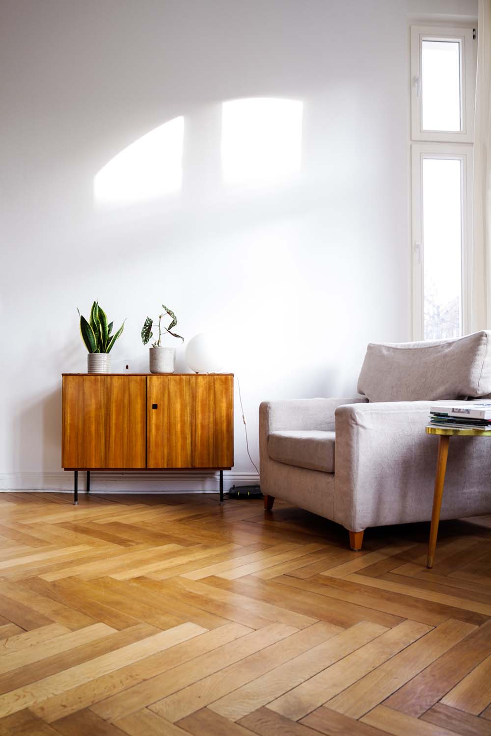 A pale wooden floor with planks arranged in a herringbone design, with an armchair and a sideboard on top. 